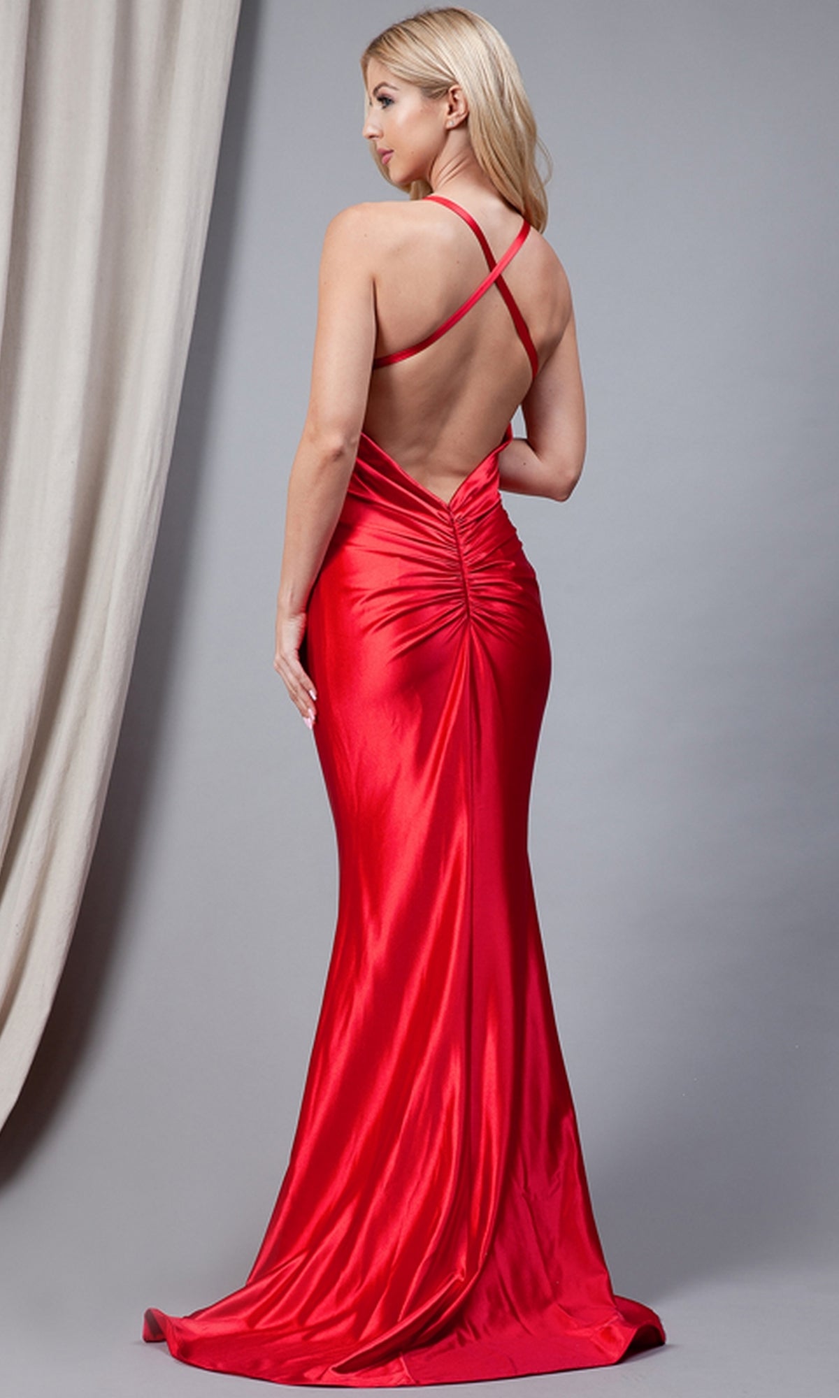 Woman in a shiny red Twist-Waist Deep V-Neck Long Prom Dress 5039 poses with her back to the camera, showing off crisscross straps, against a plain backdrop.